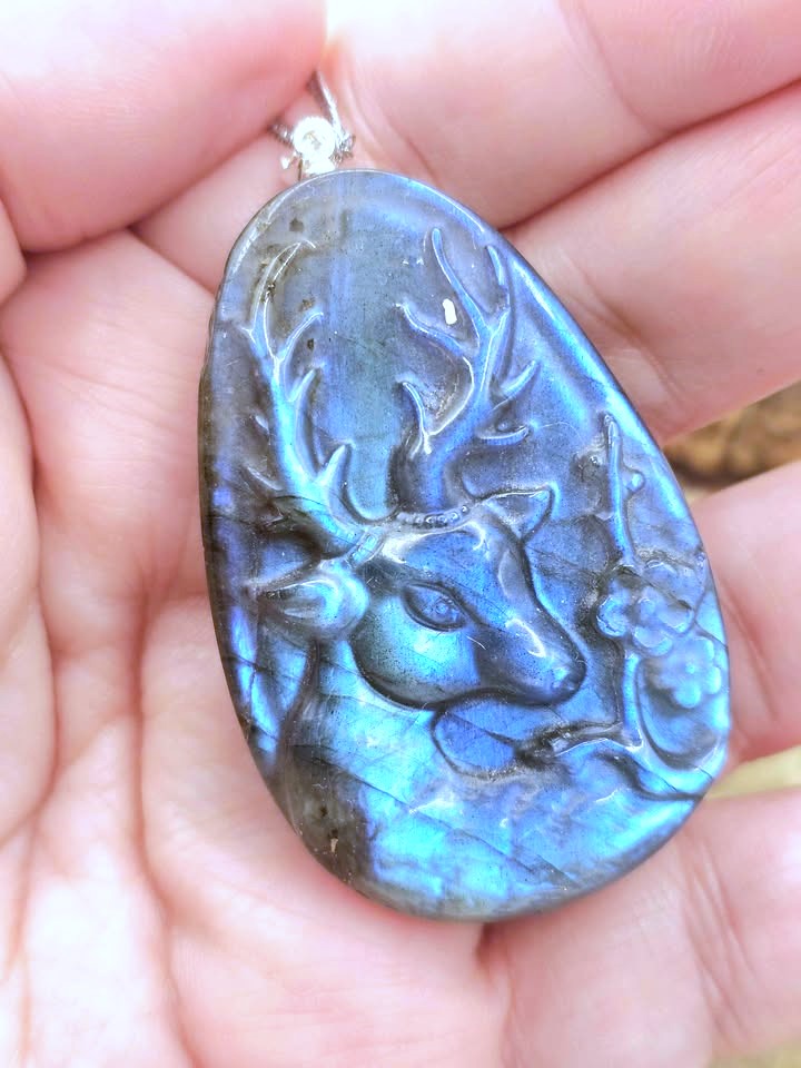 A teardrop-shaped labradorite pendant featuring a carved deer head, held in a person's hand. The pendant displays a shimmering blue and green iridescent labradorite finish with dark gray edges.