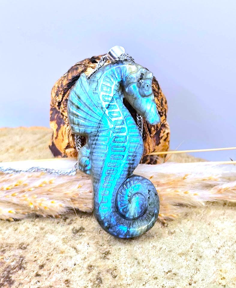 A seahorse-shaped pendant necklace with a distressed turquoise and blue finish, displayed on a piece of driftwood with a seashell in the background.