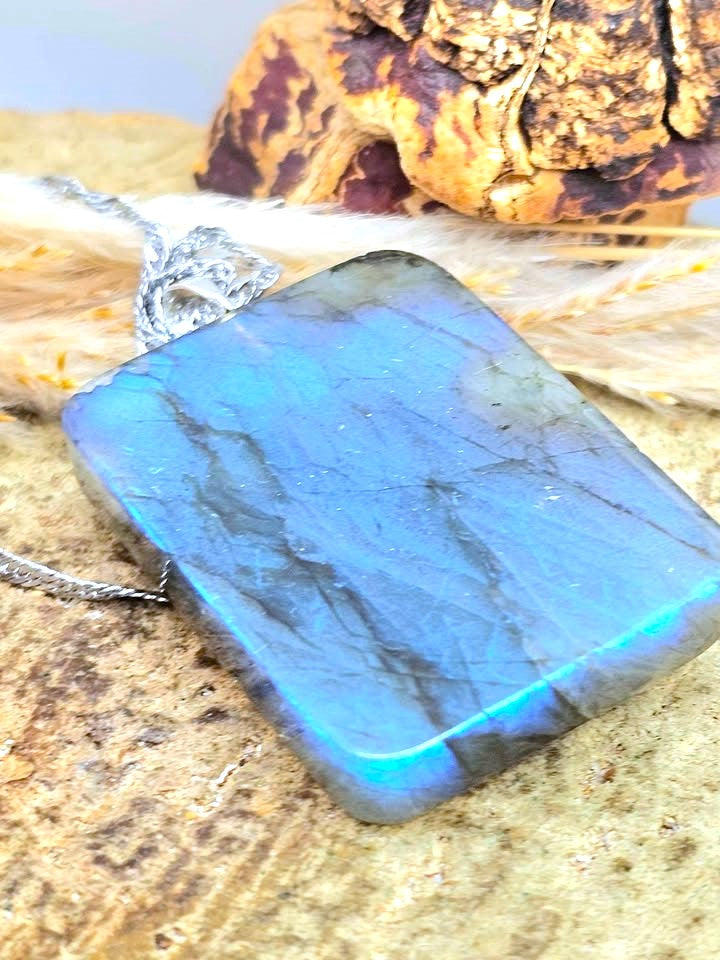 A rectangular labradorite pendant necklace with a silver chain, resting on a textured stone surface with dried grass and a decorative object in the background.