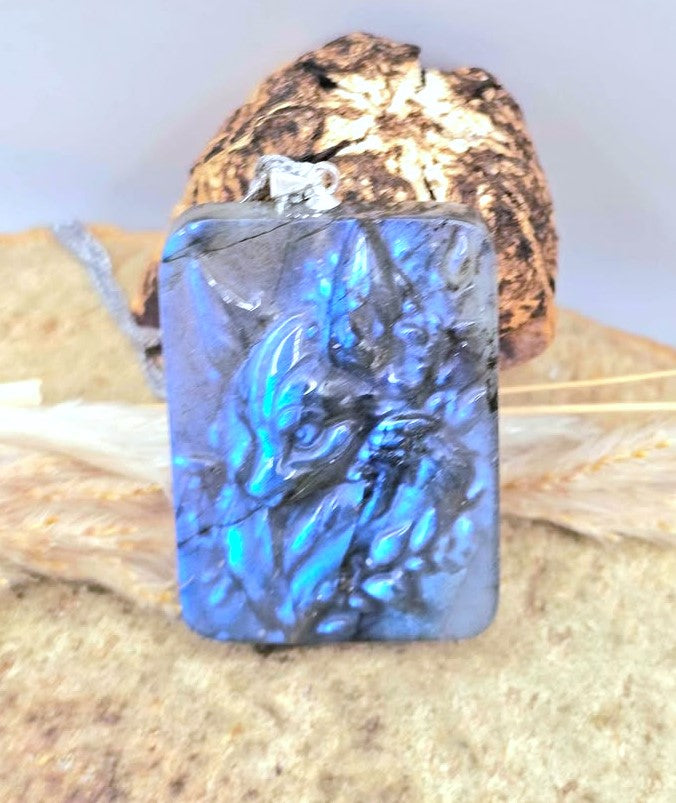 A rectangular blue labradorite pendant with a carved figure design, displayed on a textured beige surface with dried grass and a decorative rock behind it.