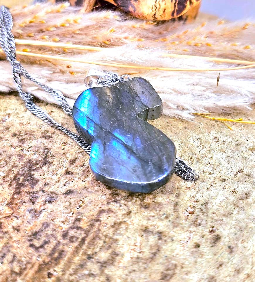 A silver pendant necklace featuring a polished labradorite stone with a vibrant blue iridescent sheen, resting on a textured stone surface with dried grass.