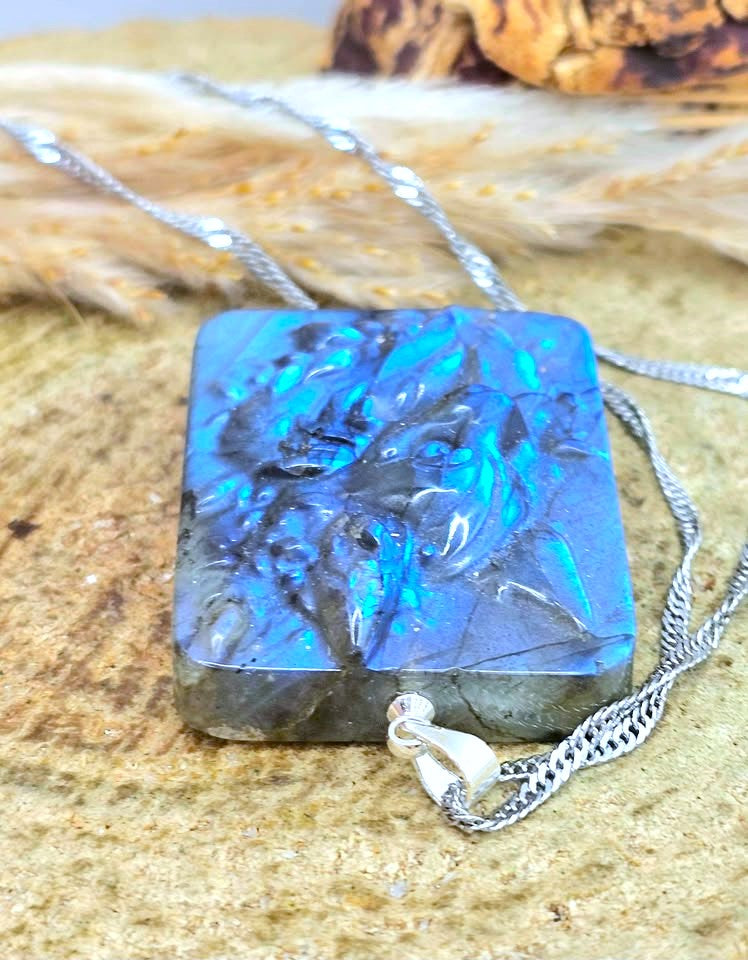 A rectangular labradorite pendant necklace with a silver chain, resting on a textured sandy surface with dried grass in the background. The pendant displays vibrant blue and green iridescent play-of-color on a dark gray base.
