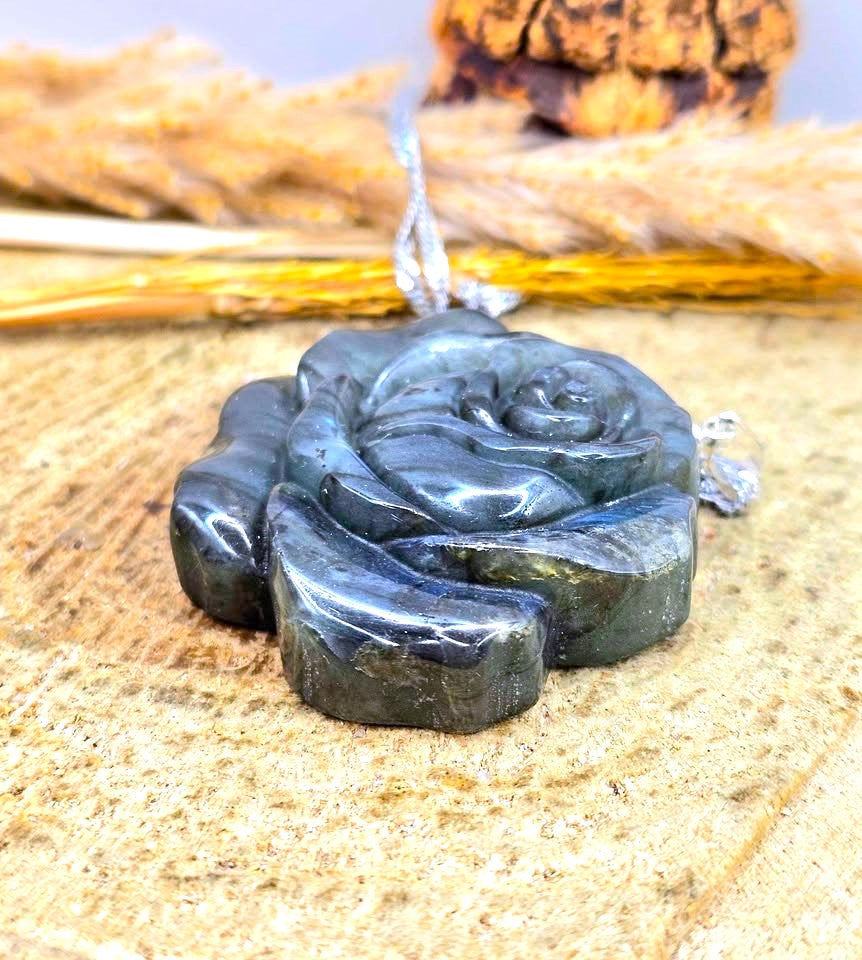 A polished dark green carved pendant shaped like a rose, suspended from a silver-toned chain, displayed on a textured beige surface with dried grass in the background.