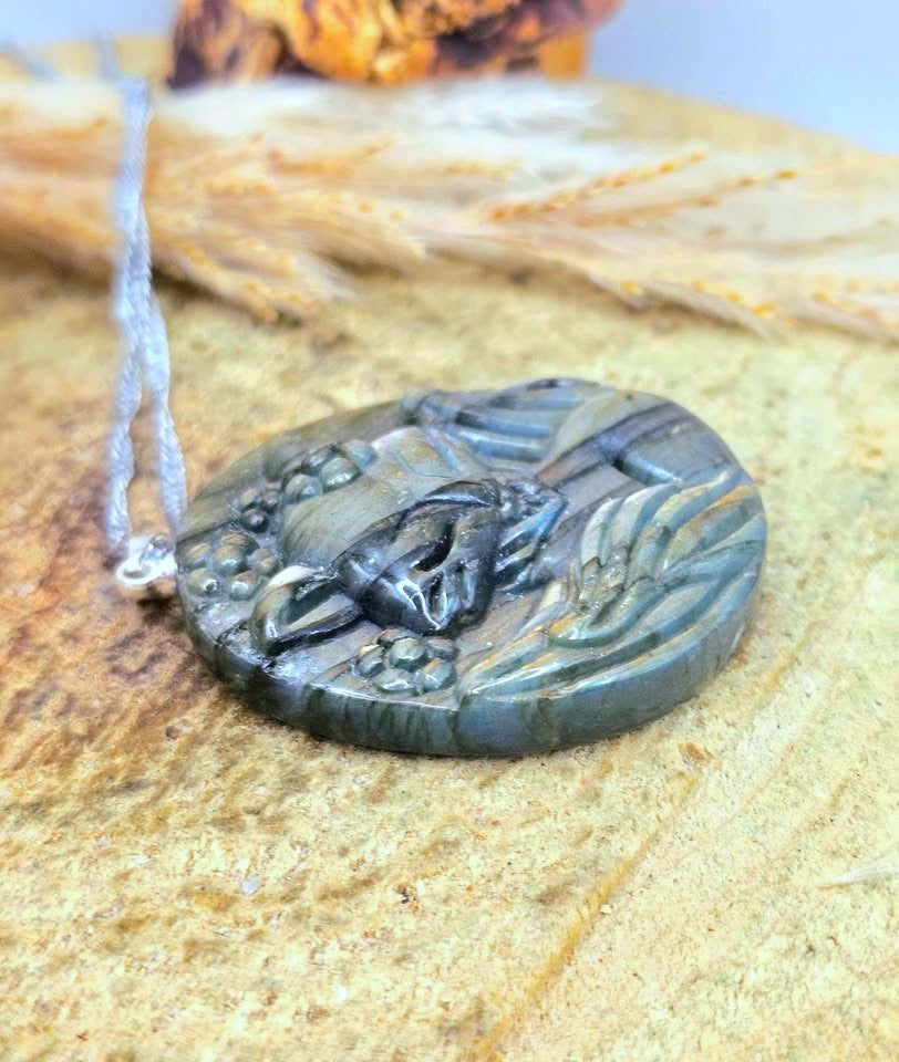 A round carved pendant necklace featuring a detailed animal carving, likely a fox or rabbit, with a silver-toned cord, displayed on a textured tan surface with dried grass in the background.
