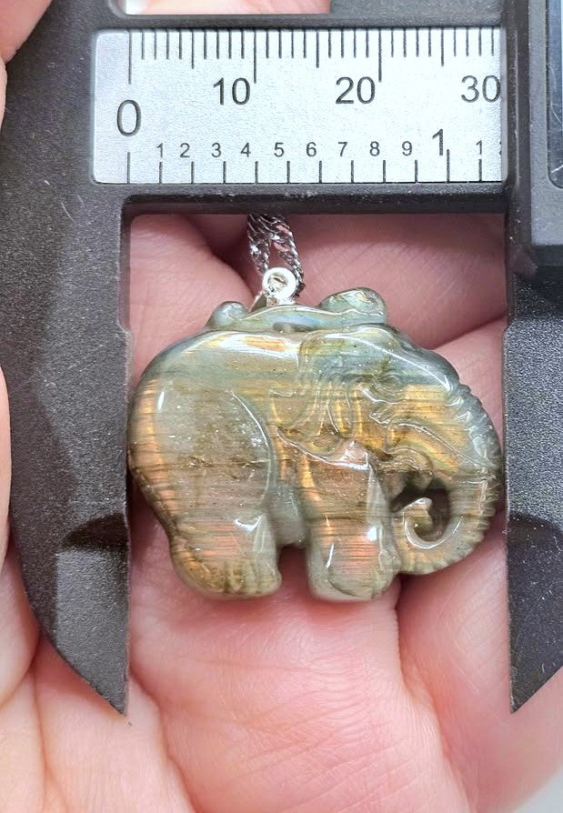 A small carved elephant pendant with a gold-toned chain, held in a person's hand under a metal caliper to show its size. The pendant has a polished, iridescent surface with shifting olive, green, and gold tones.