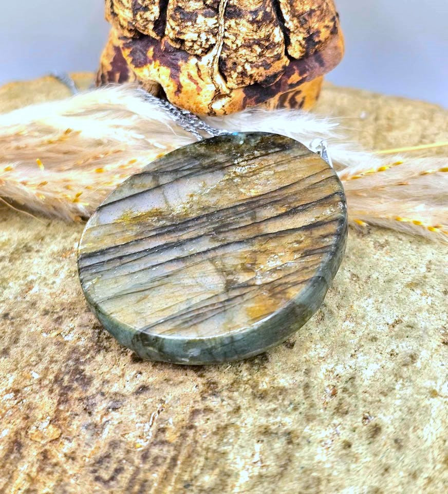 A polished round labradorite stone pendant with natural striations, resting on a textured stone surface with feathers and a decorative object in the background.