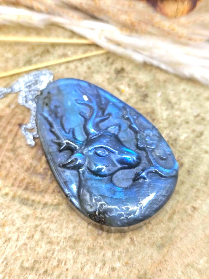 A teardrop-shaped labradorite pendant featuring a carved deer head design, showcasing a vibrant blue iridescent sheen, resting on a textured stone surface.