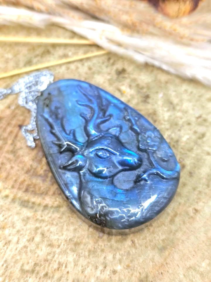 A teardrop-shaped labradorite pendant featuring a carved deer head design, showcasing a vibrant blue iridescent sheen, resting on a textured tan surface.