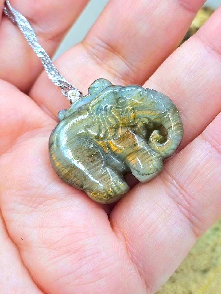 A carved elephant pendant made of polished tiger's eye stone, held in a person's hand. The pendant features natural golden-brown and olive-green banding, suspended from a silver-colored chain.