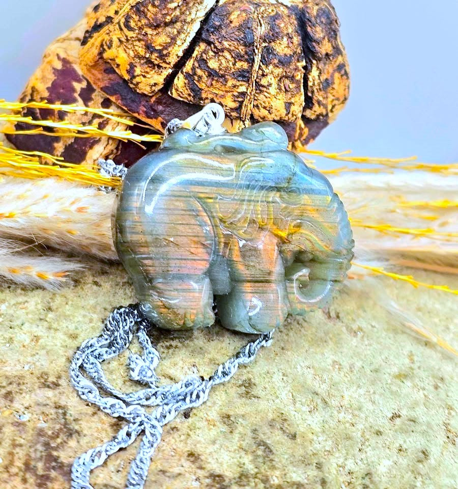 A carved elephant pendant necklace made of iridescent labradorite with blue, green, and orange bands, hanging from a silver chain on a textured stone surface with dried grass and a decorative rock in the background.