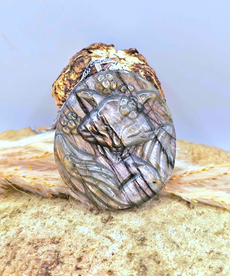 A polished oval pendant featuring a carved nine tailed fox design with small bead details, resting on a textured rock with dried grass against a light blue background.