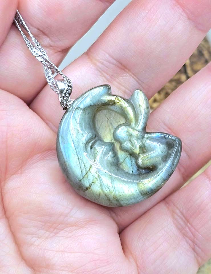A close-up of a hand holding a polished crescent moon pendant carved from labradorite, showing its characteristic green and gold iridescent play-of-color, with a silver bail and chain.