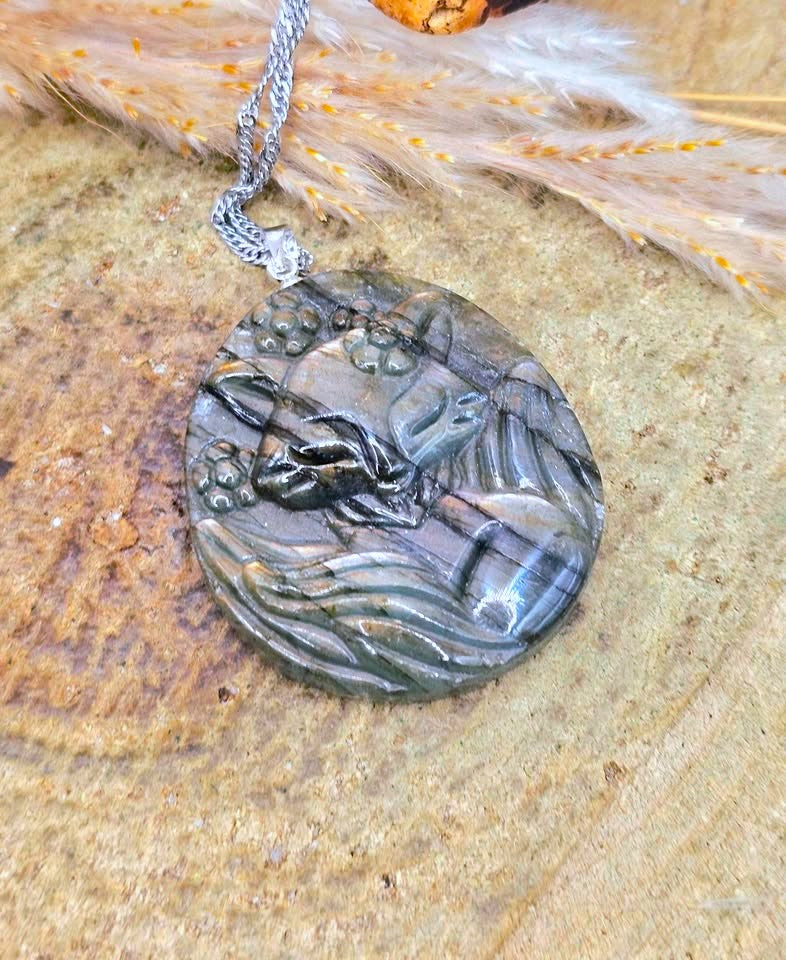 A round carved pendant necklace featuring a detailed rabbit design, crafted from a dark gray stone with natural inclusions, suspended from a silver chain and displayed on a textured wood surface with dried grass.