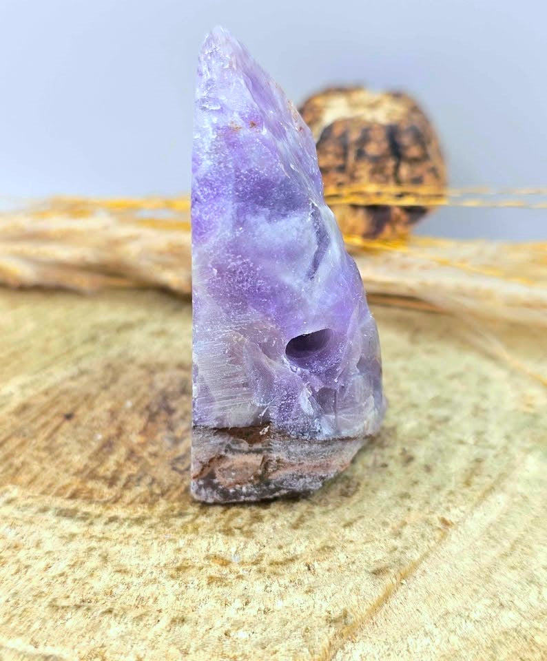 A natural purple amethyst crystal cluster with a rough, pointed tip and a dark matrix base, displayed on a wood slice with dried grass in the background.