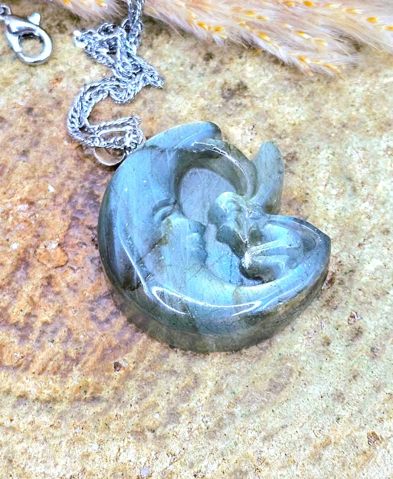 A polished labradorite pendant necklace featuring a carved figure design, resting on a textured stone surface with a silver chain and clasp.
