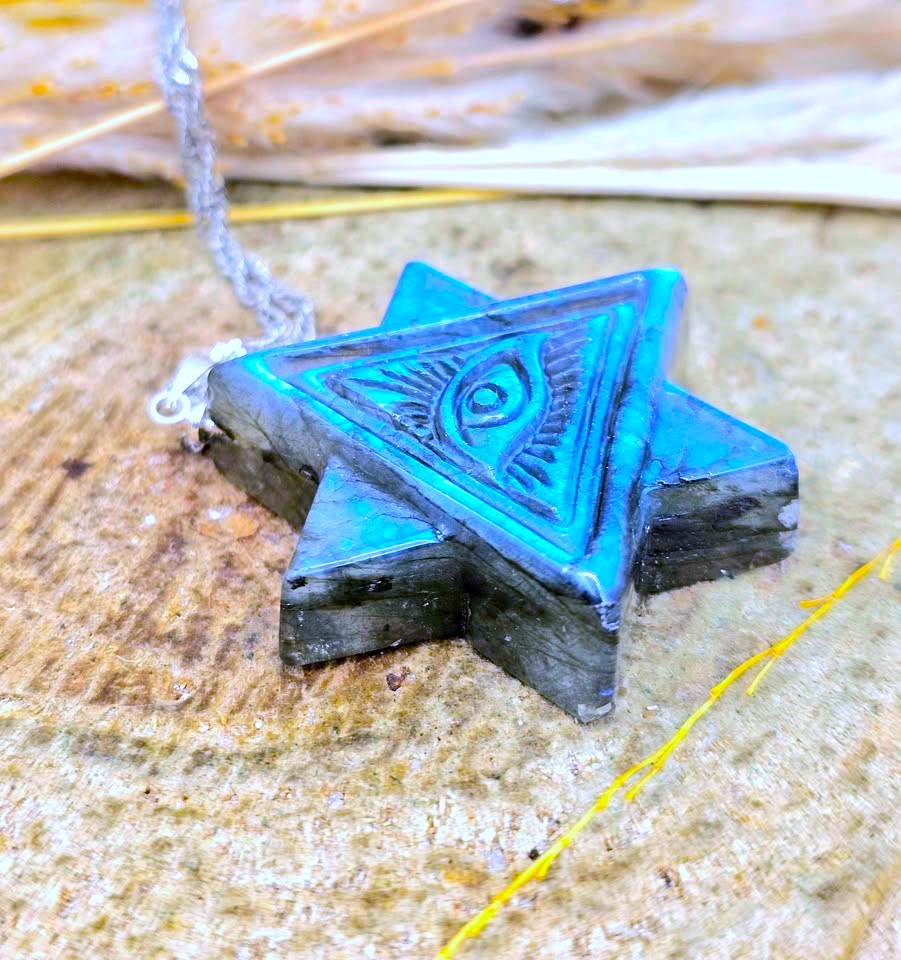 A triangular star pendant with an eye of providence symbol in the center, featuring a blue and black wood-grain finish, resting on a textured wooden surface with a silver chain.