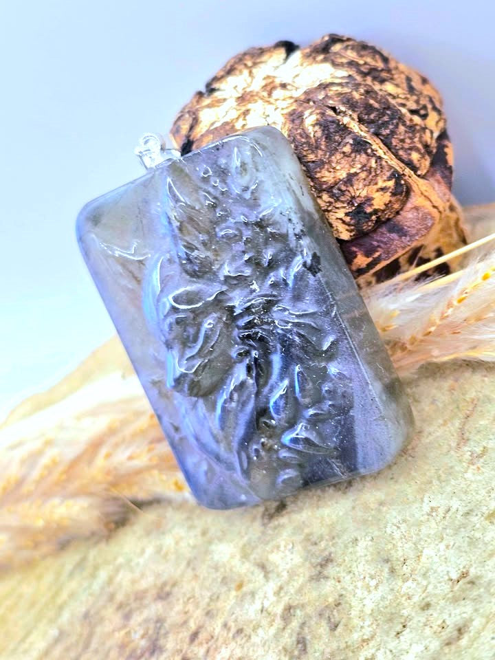 A rectangular clear resin pendant with an embossed floral or leafy design, resting on a textured beige surface with dried grass and a brown decorative object in the background.