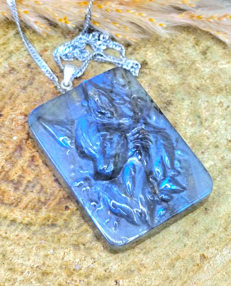 A rectangular silver pendant necklace featuring a raised relief design of a horse head, set in a polished blue labradorite stone. The pendant hangs from a silver chain and is displayed on a textured tan stone surface with dried grass in the background.