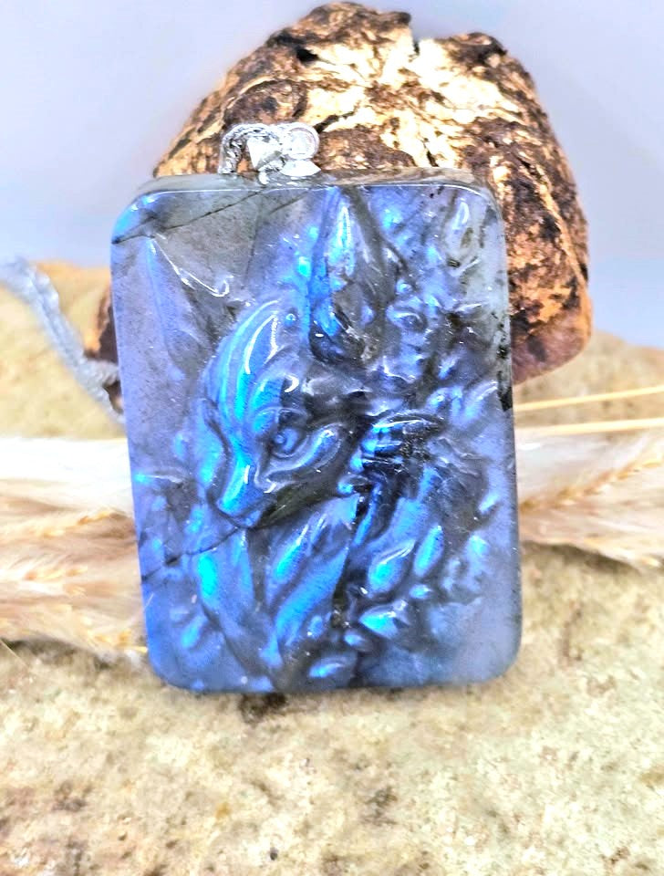 A rectangular pendant with a carved dragon design, featuring vibrant blue and black iridescent coloring, displayed on a textured sandy surface with a piece of wood in the background.