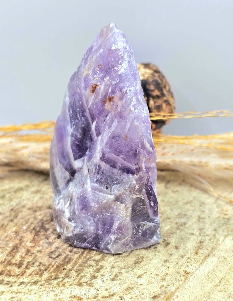 A raw purple amethyst crystal point resting on a textured beige stone surface with dried grass in the background.
