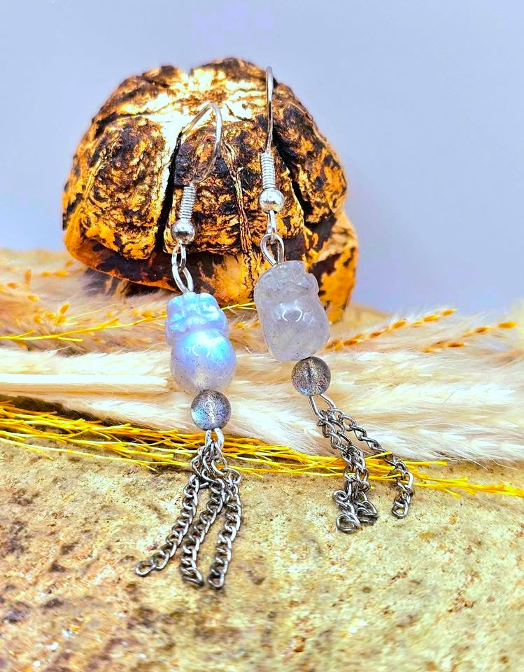 A pair of handmade dangle earrings displayed on a textured surface with dried grass and a decorative object. The earrings feature silver-toned metal chains, a blue iridescent bead, and a pale gray stone bead.