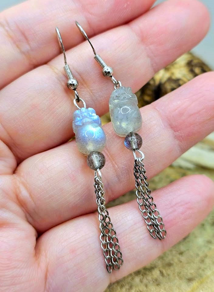 A pair of handmade dangle earrings held in a person's hand. The earrings feature silver-toned metal hooks, small silver beads, a light blue iridescent stone bead, a small carved light gray stone bead, and a long silver chain tassel.