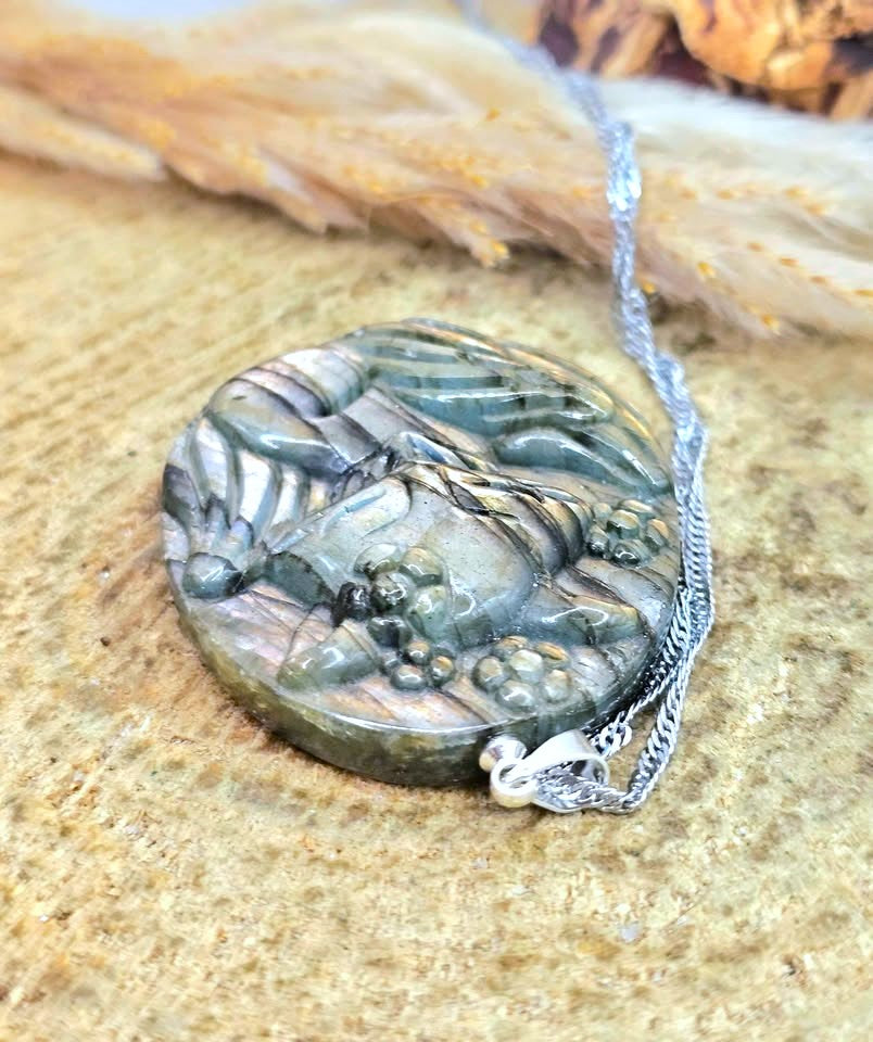 A round carved stone pendant necklace featuring a detailed animal design, attached to a silver chain, displayed on a textured sandy surface.