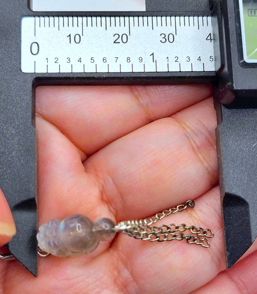 A close-up of a person's hand holding a small translucent gray glass bead with a silver chain, positioned under a metal caliper to show its size.