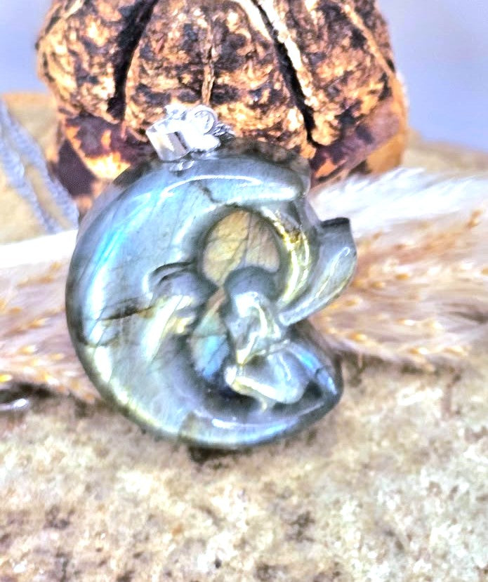 A close-up of a carved oval pendant made from iridescent labradorite stone, featuring a stylized figure design, resting on a textured surface with a brown decorative object behind it.