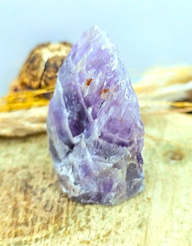 A raw purple amethyst crystal cluster with natural jagged edges, resting on a textured beige surface with dried grass and a blurred natural background.