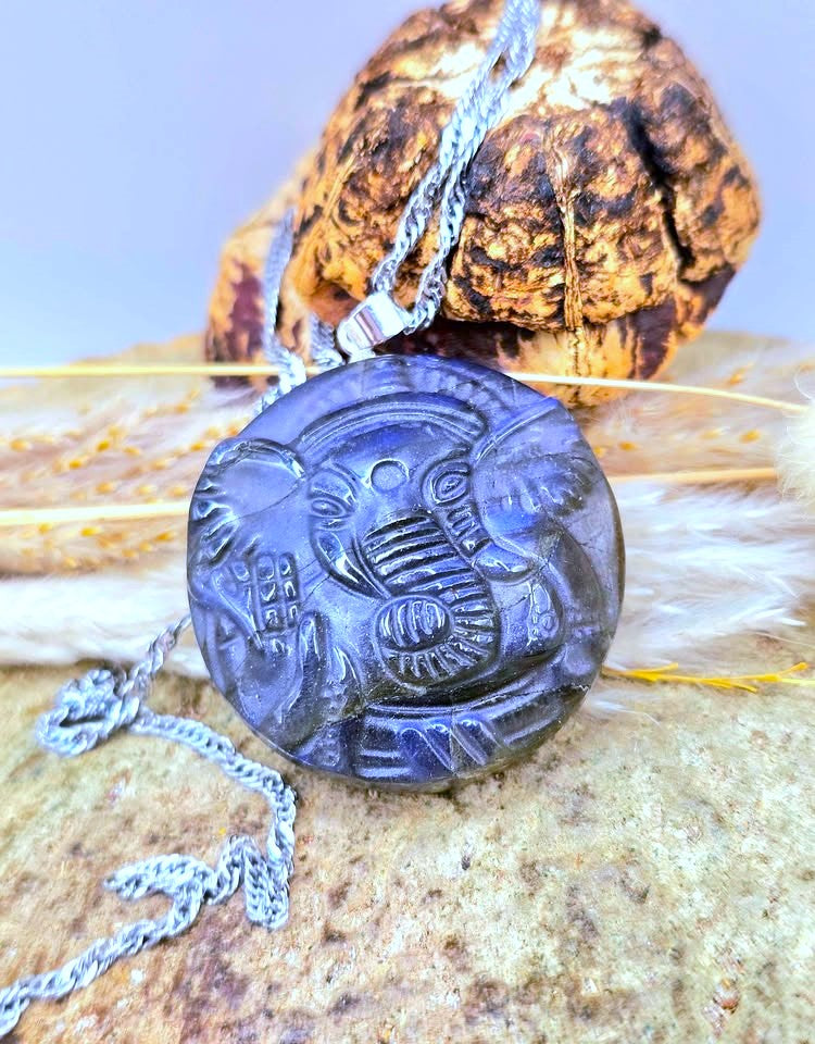 A round dark gray carved pendant necklace featuring an Egyptian cobra motif, displayed on a textured rock with a piece of wood and white feathers in the background.