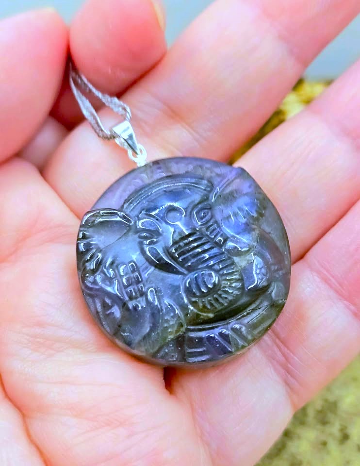 A carved oval pendant with a silver bail, held in a person's hand. The pendant appears to be made of a dark polished stone or resin with engraved details and a cool bluish-purple cast.