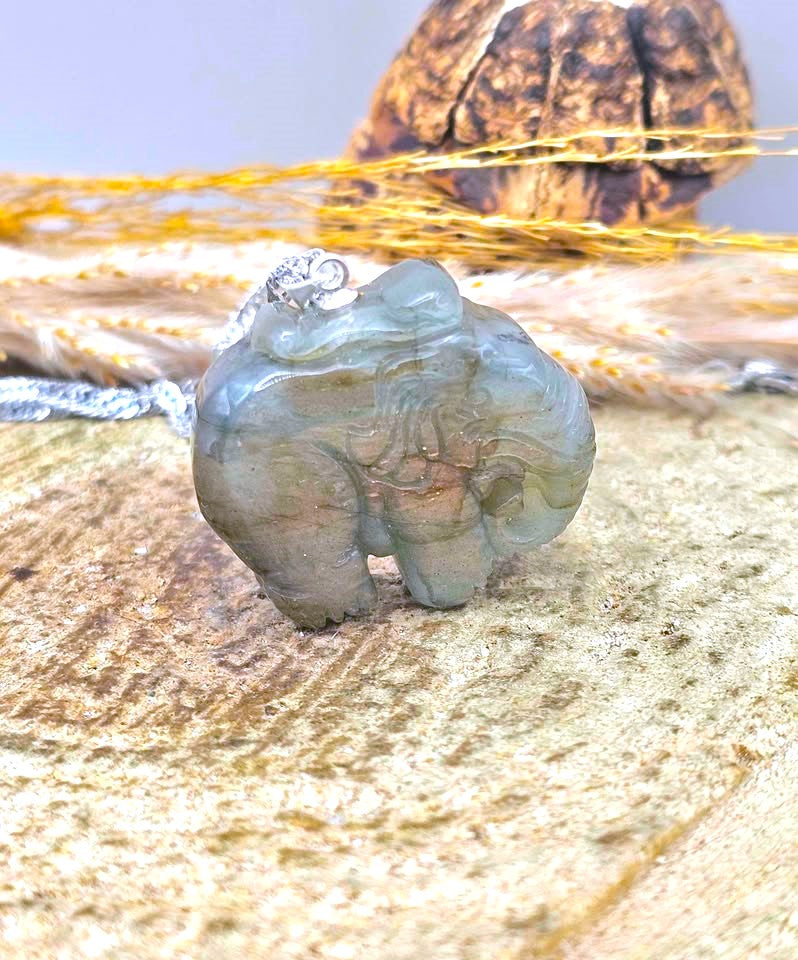 A carved elephant pendant made of pale blue-green stone with a silver-toned loop at the top, displayed on a textured surface with dried grass and a shell in the background.