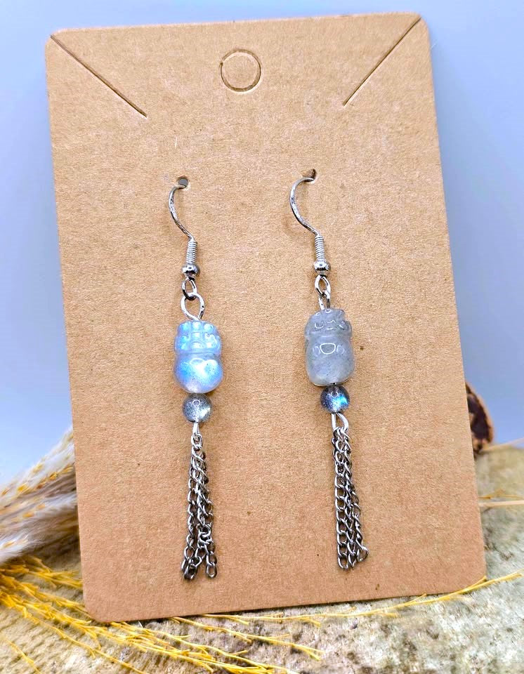 A pair of dangle earrings displayed on a brown kraft card. Each earring has a silver-tone fishhook, a small blue iridescent bead, a gray stone bead, and a dangling silver-tone chain tassel.