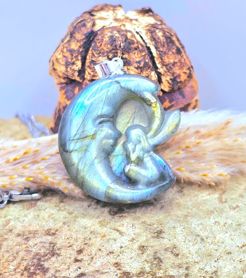 A polished labradorite crescent moon pendant with a baby figure, resting on a textured surface with a dried flower and a small silver clasp visible at the top.