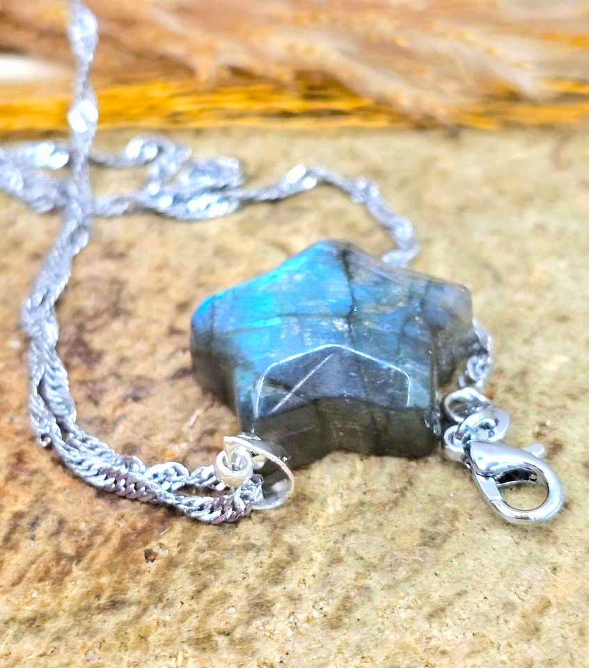 A silver chain necklace with a faceted dark gray labradorite gemstone pendant, resting on a textured tan stone surface.
