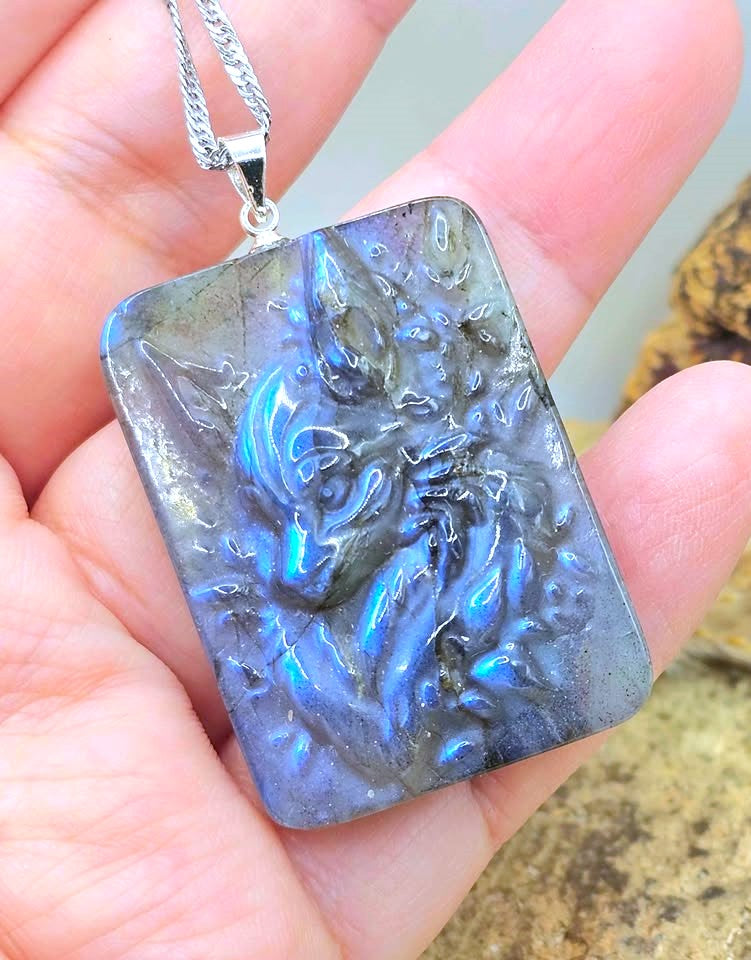 A rectangular labradorite pendant necklace with a raised dragon design, held in a person's hand. The pendant exhibits a shimmering blue and gray coloration characteristic of labradorite, suspended from a silver chain.