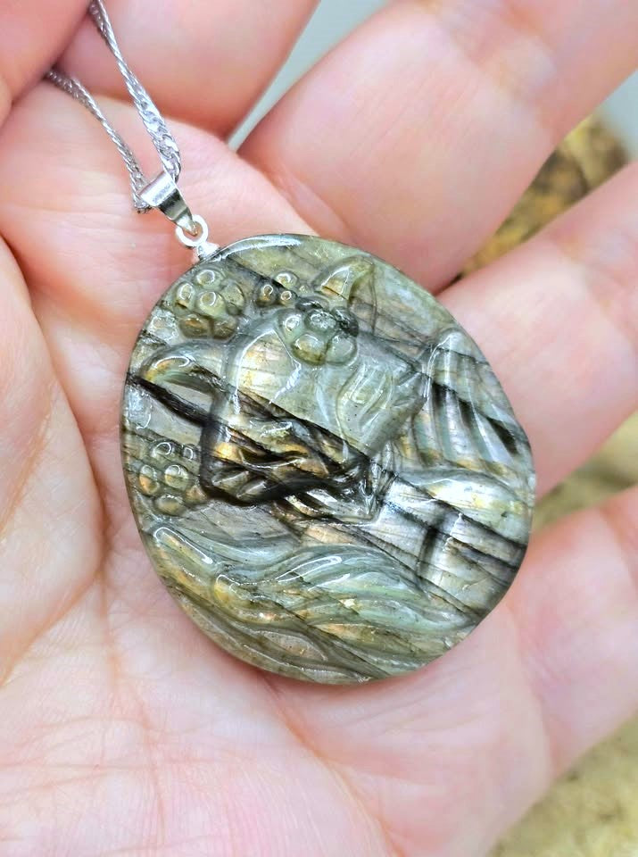 A close-up of a hand holding an oval pendant necklace. The pendant is made of a polished stone with natural gold and gray veining, featuring a carved figure design, and hangs from a silver-toned chain.