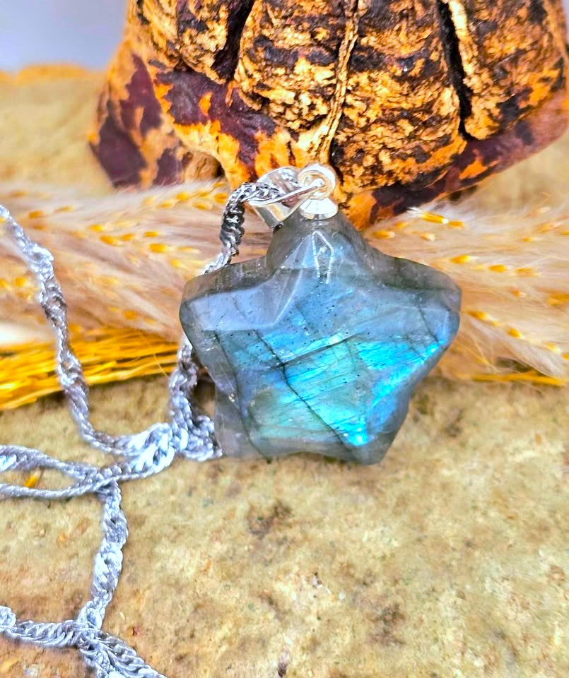 A star-shaped labradorite gemstone pendant necklace with a silver chain, displayed on a textured surface with dried grass and a decorative object in the background.
