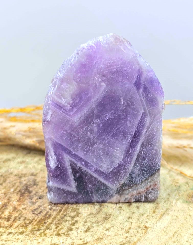 A raw purple fluorite crystal cluster with a rough, natural shape, standing upright on a textured wood slice with dried grass in the background.