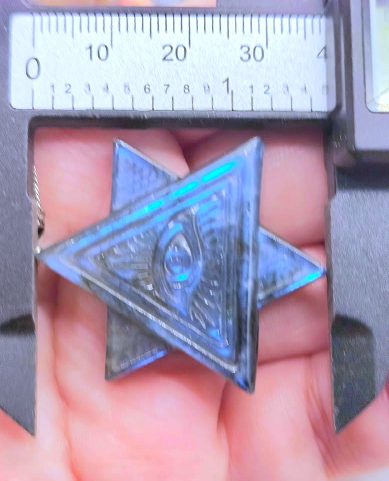 A close-up of a metallic star-shaped pendant held in a hand under a caliper, showing its blue iridescent finish and engraved eye design.