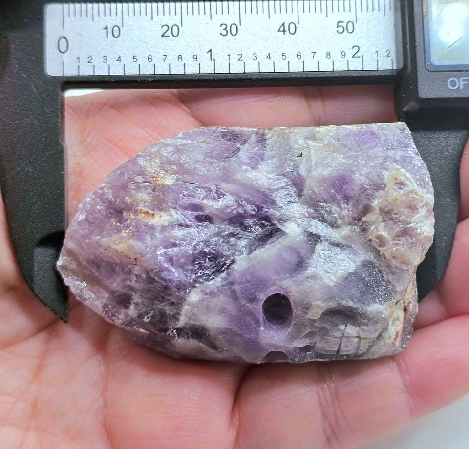 A raw purple crystal specimen held in a person's hand under a digital caliper to show its dimensions. The crystal has natural inclusions and a rough, unpolished texture.