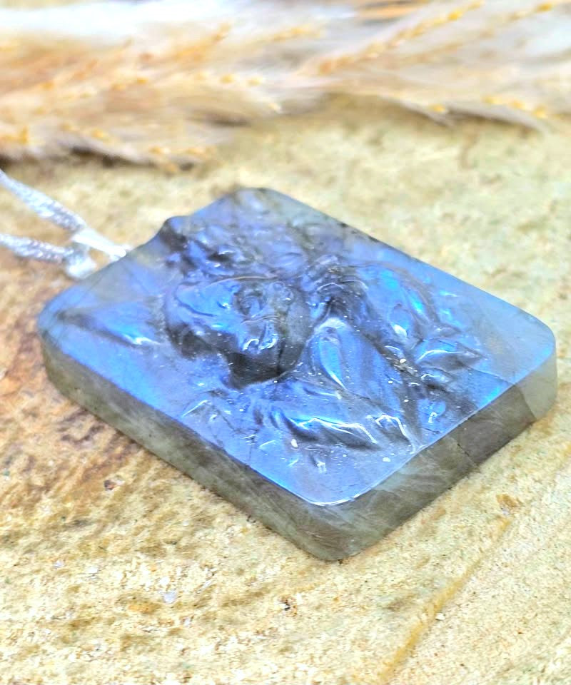 A square carved pendant necklace made of iridescent blue-gray labradorite, resting on a textured sandy surface with dried grass in the background.
