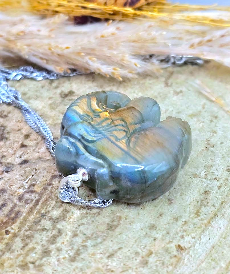 A polished labradorite gemstone pendant necklace with a silver chain, resting on a textured stone surface with dried grass in the background.