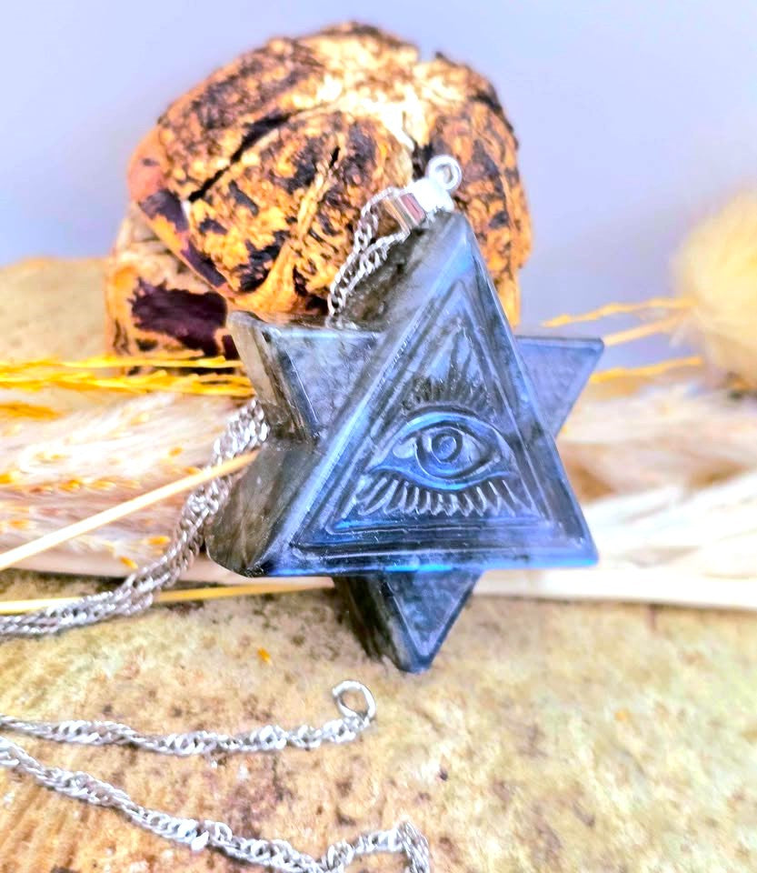 A dark blue triangular pendant necklace with an embossed eye of All Seeing symbol, suspended on a silver chain, displayed on a textured stone surface with dried grass and a dried flower in the background.