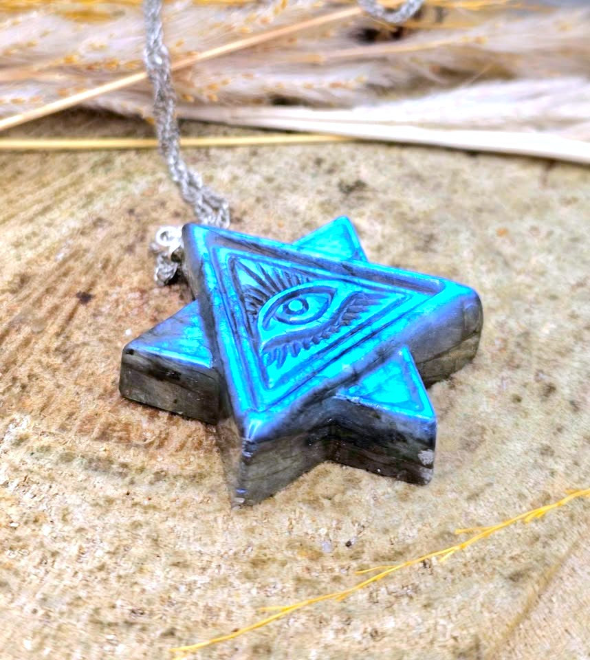A star-shaped pendant necklace with an engraved eye symbol, featuring a vibrant blue and black textured finish, hanging from a silver chain on a rustic wooden background.