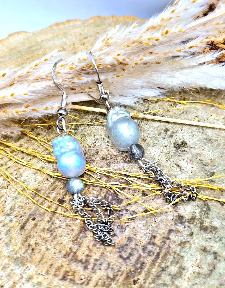 A pair of dangling labradorite pi xiu earrings displayed on a textured stone surface with dried grass. Each earring has a silver fishhook, a large iridescent blue bead, a small dark bead, and a short silver chain with a decorative dangling charm.
