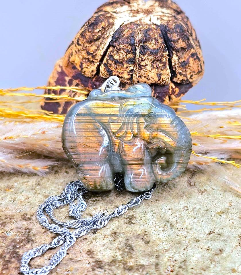 A carved elephant pendant necklace made of layered gold, orange, and gray stone, hanging from a silver chain on a textured surface with dried grass and a decorative object in the background.