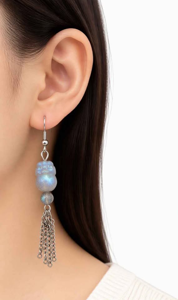 A close-up of a model's ear wearing a dangling earring with a silver-toned hook, a light blue iridescent bead, a small pearl-like bead, and a dangling silver chain.