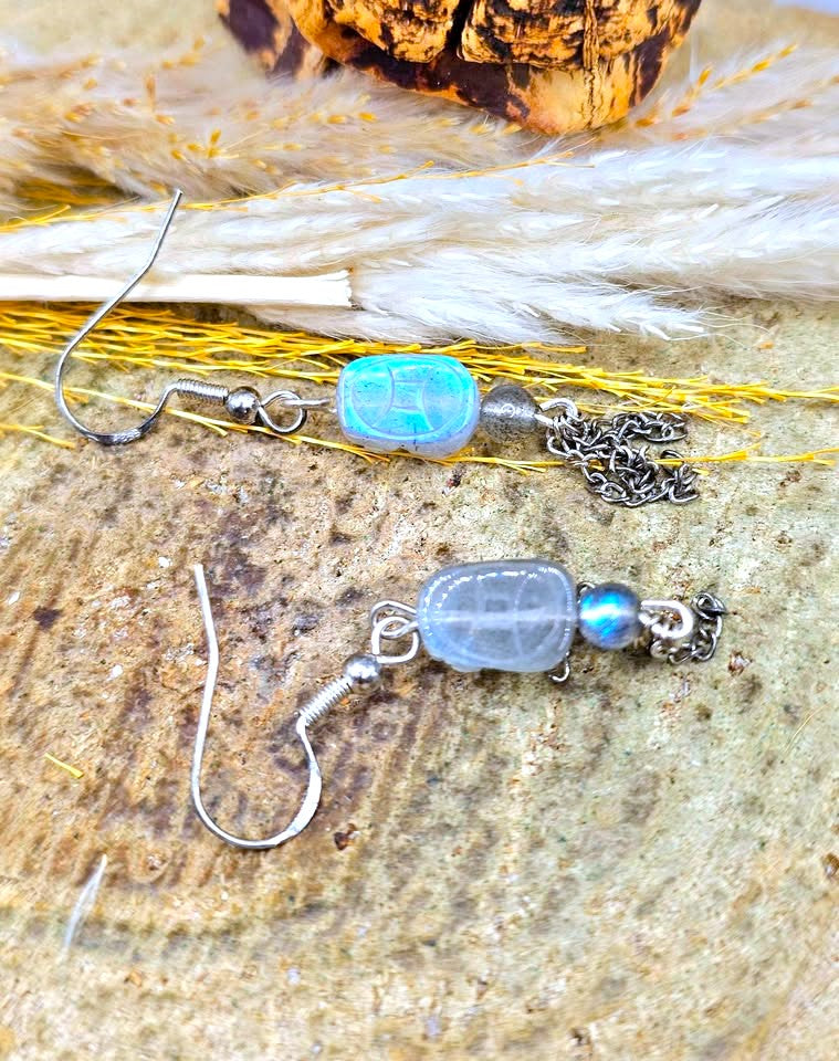 A pair of handmade dangle earrings with silver fishhook wires, featuring a light blue iridescent stone bead, a small silver chain, and a tiny blue iridescent bead, displayed on a textured stone surface with dried grass.